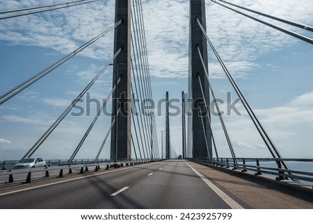 A clearskied modern suspension bridge with twin towers and cables, calm seas on either side. An empty lane, white car seen, possibly the Oresund Bridge linking Copenhagen to Malmo.