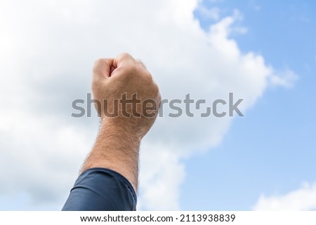 Clenched Fist In The Air.male hand raises clenched fist of solidarity. protester holding first up hand over bue sky. hand rising up to sky.Man clenched fist raised in triumph or defiance, against sky