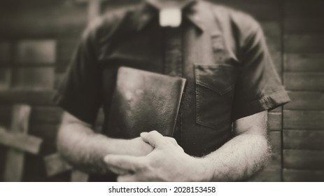 Clergyman, Reverend Or Priest Wearing A Clerical Collar And Clutching A Bible. Preacher Preaching The Gospel In Front Of An Old Rustic Rural Church Or Cemetery.