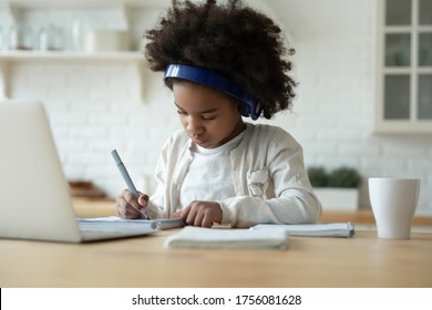 Clever African schoolgirl excellent pupil sit in kitchen do homework wears headphones writing on workbook listen audio lesson use laptop. Videocall, e-study with tutor distantly, homeschooling concept