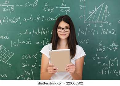 Clever attractive young female college student wearing glasses and holding a tablet computer in her hands standing in front of a chalkboard covered in equations in maths class smiling at the camera