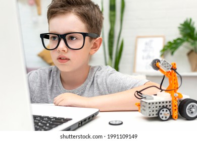 Clever boy in eyeglasses builds and programs a robot car using a laptop at home. The child is learning coding and programming. Robotics, Science, Mathematics, Engineering, Technology. STEM education.