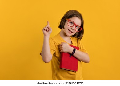 Clever Child School Girl Pointing Up While Reading Book. Little Girl 8-10 Years Old In Yellow T-shirt And Red Glasses On Yellow Background Hold Red Textbook. Smart Kid Have Excellent Idea