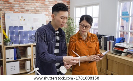 Clever creative architect team. Qualified skilled smart asian woman and man engineers feeling confident while holding tablet and looking at lovely model of windmill turbine. two colleagues in office