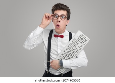 Clever excited man in bowtie and shirt looking at camera through spectacles in amazement while holding computer keyboard on gray background - Shutterstock ID 2074905586