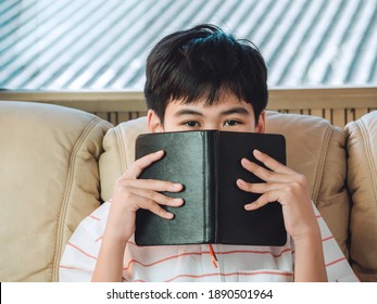 Clever Little Boy Hiding Face Behind The Black Cover Notepad With Eyes Wide Open. Kid Is Playing Hide And Sick. Genius Concept