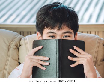 Clever Little Boy Hiding Face Behind The Black Cover Notepad With Eyes Wide Open. Kid Is Playing Hide And Sick. Genius Concept