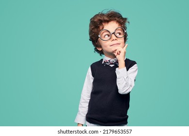 Clever little student in school uniform and glasses touching face and looking away while pondering during studies against turquoise background - Shutterstock ID 2179653557
