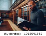 Clever professional lawyer spending time on learning laws in juridical literature in public library, concentrated man journalist reading nonfiction book analyzing information standing news bookshelf