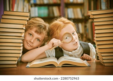 Clever schoolchildren in neat uniform sit in a school library among piles of books studying curiously an encyclopedia with a magnifying glass. Modern education and children.