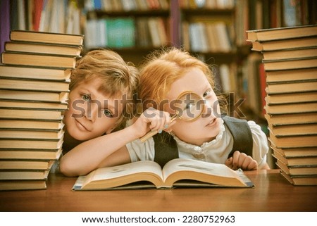 Clever schoolchildren in neat uniform sit in a school library among piles of books studying curiously an encyclopedia with a magnifying glass. Modern education and children.