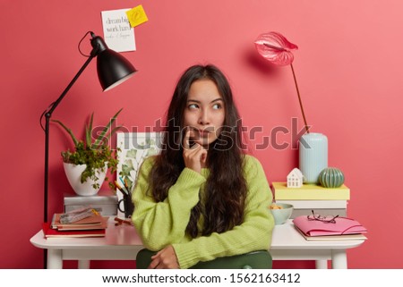 Clever thoughtful female student looks away, contemplates something, thinks about idea of making sketch for watch, prepares project, wears green sweater, poses against workplace over pink wall