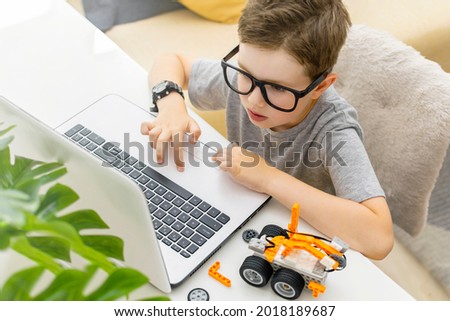 Clever young schoolboy in glasses builds and programs a robotic vehicle codes an electronic toy at home. Child using laptop. STEM education and programming lesson concept. Top view.