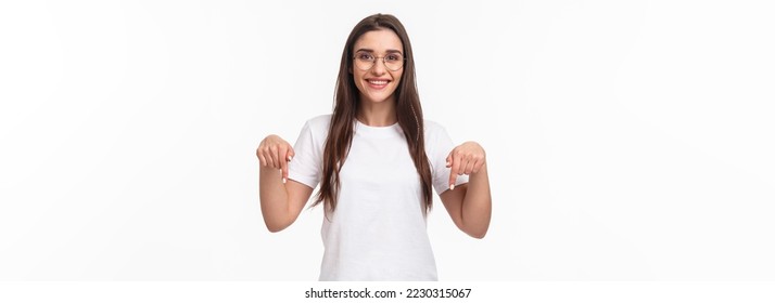 Click here. Portrait of brunette gorgeous young woman in glasses, pointing fingers down to show advertisement, smiling pleased, suggest visit link, recommend download or subscribe, white background.