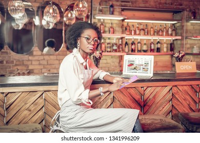 Clicking on table at bar. Serious pretty Afro-American curly-haired bar worker in glasses and white shirt with apron clicking on tablet while laptop is on bar counter - Powered by Shutterstock