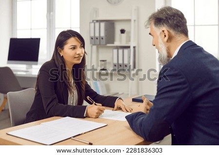 Client and business agent in the office. Young woman sitting at an office desk signs a paper contract after listening to the explanations of a professional loan broker or financial adviser