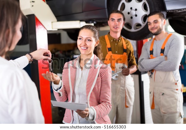 Client at a car dealership giving keys from\
her car to vehicle repair crew manager. Confident smiling mechanics\
are standing in a\
background