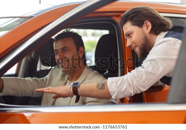 Client\
of car dealership sitting inside vehicle, smiling and testing car.\
Bearded manager standing near auto and showing car with hand.\
Concept of purchase of automobile in car\
center.