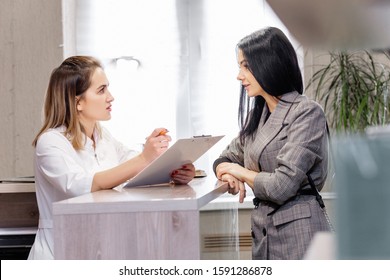 Client And Doctor Discuss Cosmetic Procedure In Beauty Clinic. Young Stylish Brunette Girl Makes Appointment With Beautician At Reception With Administrator. Concept Anti-aging Treatment And Injection