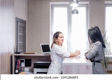 Client And Doctor Discuss Cosmetic Procedure In Beauty Clinic. Young Stylish Brunette Girl Makes Appointment With Beautician At Reception With Administrator. Concept Anti-aging Treatment And Injection