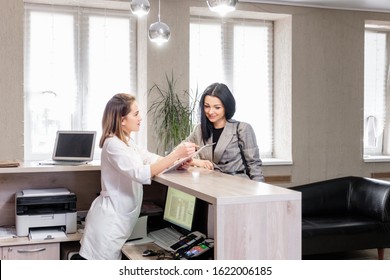 Client And Doctor Discuss Cosmetic Procedure In Beauty Clinic. Young Stylish Brunette Girl Makes Appointment With Beautician At Reception With Administrator. Concept Anti-aging Treatment And Injection