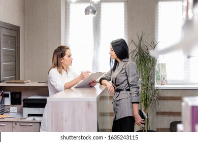 Client And Doctor Discuss Cosmetic Procedure In Beauty Clinic. Young Stylish Brunette Girl Makes Appointment With Beautician At Reception With Administrator. Concept Anti-aging Treatment And Injection