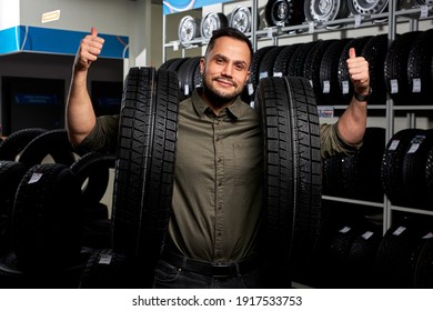 Client Guy Stands With Two Tires By Rack Of Tires, He Made Choice, Buy The Best Ones In Auto Service Shop, Showing Thumbs Up At Camera, Happy. Portrait