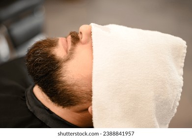 A client is lying on a chair in a barbershop with his beard wrapped in a towel. A towel covers the mans eyes. - Powered by Shutterstock