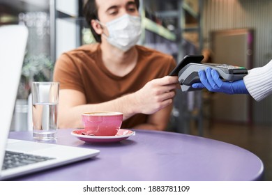 Client Mane Wearing Protective Mask Holding Contactless Credit Card At His Smartphone Near Payment Terminal While Buying Coffee In Modern Cafe. Pandemic Concept