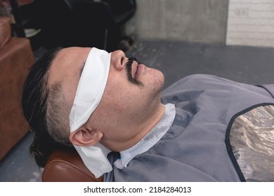 A client with a mustache at a barber shop with his eyes covered with tissue paper and neck with paper strip and a barbers cape. Getting ready for a shave. - Powered by Shutterstock