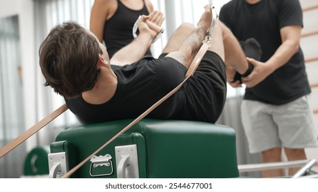 Client performing a Pilates reformer workout using straps, guided by instructor supporting. Healthy man working out pilates and hanging pulling up on the cadillac for developing upper body. Habituate. - Powered by Shutterstock