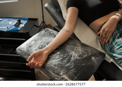 Client resting an arm on a plastic wrapped support during a tattoo session, surrounded by tattoo equipment and tools in a vibrant studio atmosphere