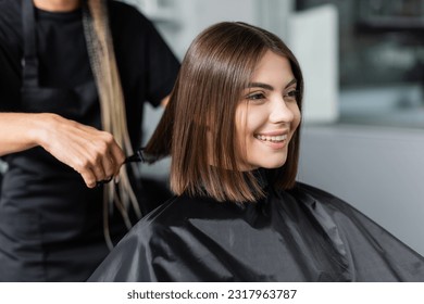 client satisfaction, cheerful woman with short brunette hair sitting in hairdressing cape in beauty salon, getting haircut by professional hairdresser, beauty salon