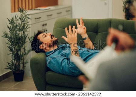 A client with trauma and problems is lying down on a sofa in a psychotherapist's office and discussing during the session. A man is talking about problems while a psychotherapist is taking notes.