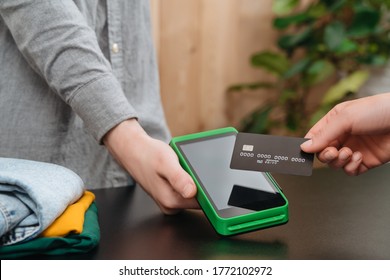 Client's hand holding bank credit card near nfc payment terminal in hand of cashier in clothing store. Customer makes purchases with credit card. Contactless, mobile, express payment, nfc concept  - Shutterstock ID 1772102972