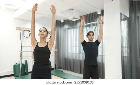 Clients practicing a standing stretch using Wunda Chair equipment during a Pilates workout for improving flexibility, leg strength at gym. Attractive group stretching and bending down. Habituate. - Powered by Shutterstock