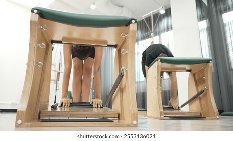 Clients practicing a standing stretch using Wunda Chair equipment during a Pilates workout for improving flexibility, leg strength at gym. Attractive group stretching and bending down. Habituate. - Powered by Shutterstock