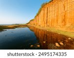 A cliff of beautiful columnar basalt, a popular tourist attraction in Xiyu, Penghu County, Taiwan, bathed in warm sunlight on a sunny summer morning and reflected in the water of a pond under blue sky