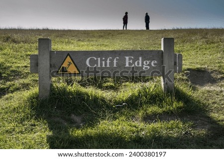 Cliff Edge old wooden sign in Beachy Head chalk headland near Eastbourne, East Sussex, South England, UK. Two people in the distance close to the edge