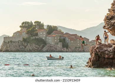 Cliff Jumping Into The Sea With Famous Sveti Stefan Island At Background In Montenegro. Group Of Friends Jumps Into The Water From The Cliff At Sunny Summer Day In Adriatic Sea, Budva, Montenegro.