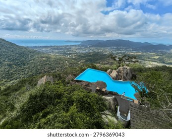 The Cliff Sky Pool is a large infinity pool perched on a rocky mountain cliff, offering a panoramic view of a coastal city and the ocean below under a cloudy sky.