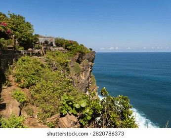 Cliffside vegetation frames ocean's vast blue expanse. Rocky cliffside grows lush green under clear sky. White foam meets deep blue waters below