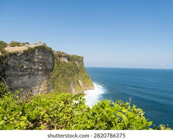 Cliffside vegetation frames ocean's vast blue expanse. Rocky cliffside grows lush green under clear sky. White foam meets deep blue waters below