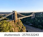 Clifton Suspension Bridge spans the Avon Gorge in Bristol, connecting clifftops with an impressive design. The sun sets, illuminating the bridge against lush greenery and serene water.