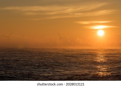 Climate Change And Global Warming. Misty Sunrise Over Offshore Wind Farm. Beautiful Orange Sky With Clean Energy Wind Turbines. Mist And Low Cloud On Sea Horizon With Hot Sun Reflecting Off The Water.