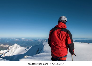 Climber In A Red Jacket Stands On Top Of A Mountain Against A Blue Sky
