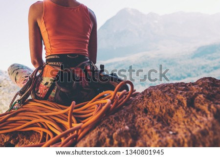 Climber sits near the rope and prepares to overcome the route. Slender girl holds climbing equipment. A woman on the background of beautiful mountains. Rest after climbing.