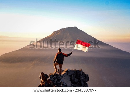 a climber stands holding a flag overlooking Mount Sumbing, Indonesia