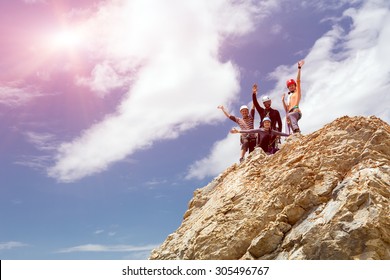 Climbers on summit. Happy people embracing raising hands up on pointed rocky cliff blue sky background 