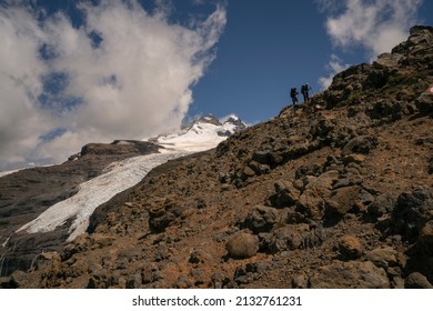 Climbing Tronador Hill. View Of Two Hikers Silhouette Trekking Along The Rocky Mountain. Glacier Castaño Overo And The Mountain Peak In The Background.